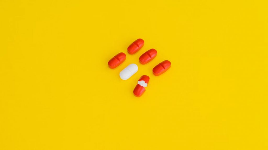 Red capsules and a white tablet arranged on a bright yellow surface, symbolizing healthcare.
